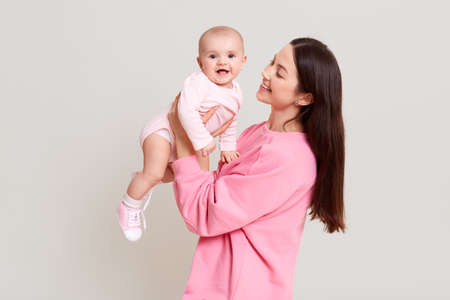 Young cheerful beautiful woman holding baby boy in her hands and looking at her with love, excited toddler girl wearing bodysuit looks at camera, family posing isolated over white background.の写真素材