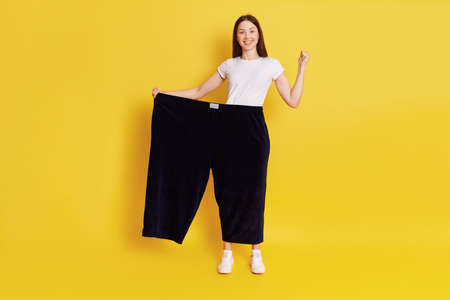Happy dark haired female wearing white t shirt and black pants huge size, brags about your result, looking at camera with satisfied expression and clenching fist with happiness.の写真素材
