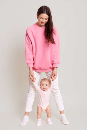 Mother teaching her little baby to walk isolated over white background, dark haired woman wearing...