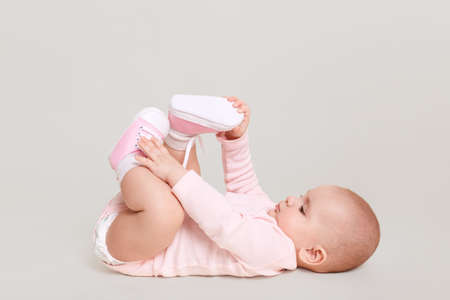 Baby lying on floor and playing with her feet, charming infant wearing pink bodysuit and shocks indoor, cute kid isolated over white background.の写真素材