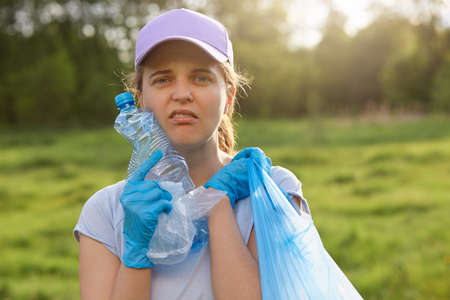 Portrait of young female volunteer posing outdoor, collecting garbage, woman in casual clothing picking up waste in field, land pollution, environmental problem.の写真素材