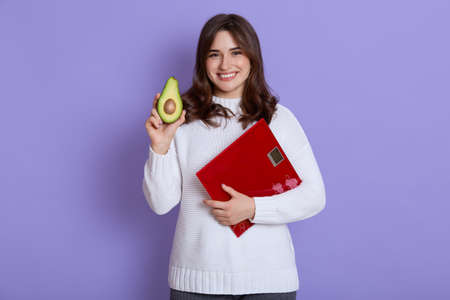 Happy smiling skinny female wearing white sweater holding floor scales and half of avocado in hands, looks at camera with positive expression, healthy lifestyle, isolated over lilac background.の写真素材