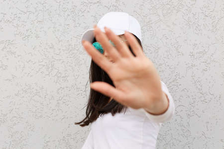 Positive dark haired female showing palm to camera, stop gesture, happy expression, wearing white shier and baseball cap, shirt and sunglasses, posing isolated over light wall outdoor.の写真素材