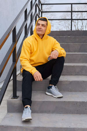 Young handsome brunette man sitting on stairs outdoors, looking away with pensive dreaming...