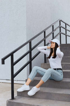 Pensive woman looking faraway with serious and thoughtful expression, keeping hands on her head, stylish lady wearing jeans, white shirt and baseball cap, model posing on stairs outdoor.の写真素材