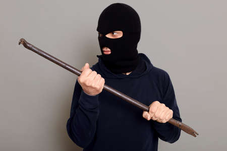 Man thief with handy crowbar looks away, wants to break open door or lock, dresses in balaclava and turtleneck, dangerous bandit commits a crime, posing isolated over gray background.の写真素材