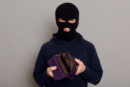 Upset burglar dressed in black turtleneck and balaclava holding empty wallet, feeling frustrated, looking at camera with pouting lips, being unhappy with theft, isolated over gray background.の写真素材