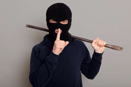 Young guy robber man dressed in a black hoodie with masked face, holds iron crowbar in his hands, shows silence gesture, keeping fore finger near his lips, isolated on gray backgroundの写真素材