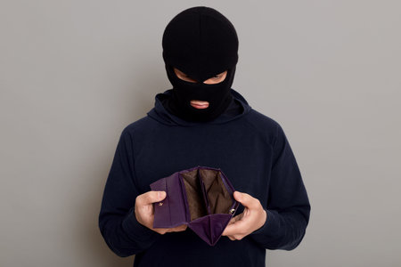 Frustrated guy thief dressed in a black hoodie and a robbery mask, holding a stolen open empty...