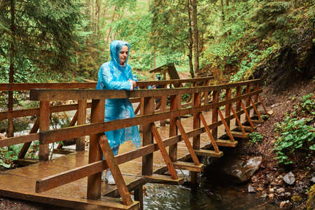 Young beautiful girl, dressed in blue raincoat, stands on wooden bridge, looking far away, enjoying nature, admires the beauty of the forest, rainy day. Outdoor full length portrait.の写真素材