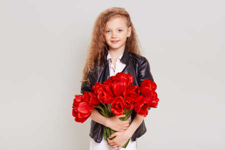 Pretty schoolgirl with wavy blonde hair wearing leather jacket standing with red tulips in hands and looking directly at camera with cute expression, isolated over gray background.の写真素材