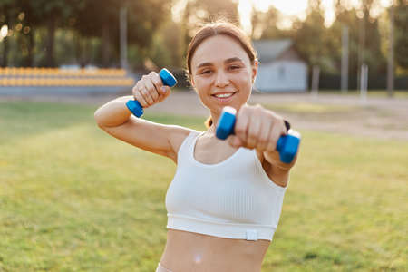 Portrait of happy satisfied smiling female wearing white top holding out dumbbell to camera, training arms, doing exercises for biceps and triceps, expressing positive emotions while doing sport.の写真素材