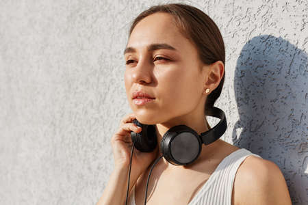 Portrait of dreamy brunette woman wearing white top and having headphones over neck, looking away...