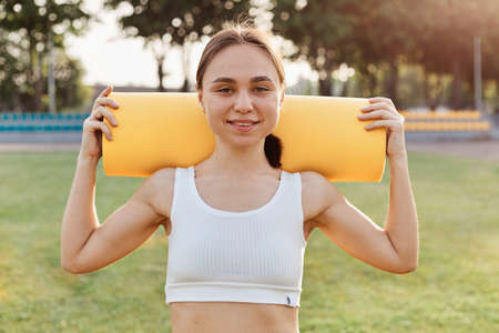 Good looking brunette female wearing white to holding yellow karemat on shoulders, looking smiling directly at camera, training outdoor in stadium in sunny day.の写真素材
