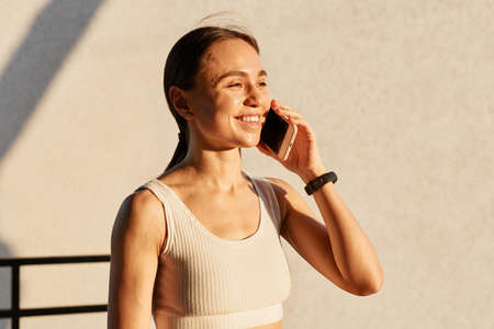 Portrait of sporty girl talking on smartphone near gray wall, looking smiling in distance, wearing white top, hearing pleasant news, expressing happiness.の写真素材
