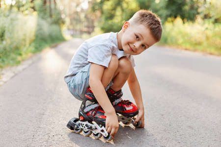 Boy in roller skating sitting on the road, looking at camera with smile, wearing white t-shirt and...