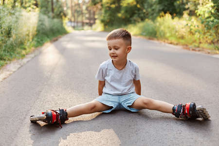 Outdoor shot of happy little boy sitting in twine on asphalt road in roller skating, looking smiling aside, learning to roller skating in summer park.の写真素材