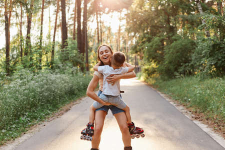 Outdoor shot of mother with little boy kid roller skating in summer park, mother holding child in hands, hugging and smiling with happiness, enjoying spending time with son.の写真素材