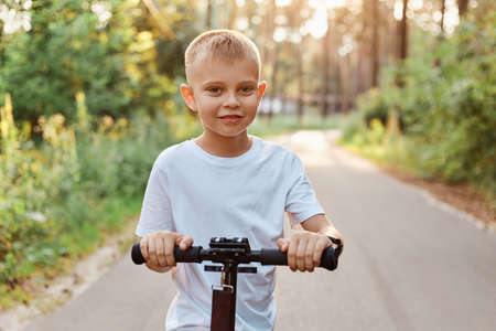Outdoor shot of happy little blond boy playing with his scooter at asphalt road in summer park, cute kid wearing white casual t shirt, happy childhood, healthy lifestyle.の写真素材