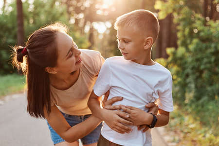 Outdoor shot of attractive woman mother hugging her little son wearing in white t shirt, mommy looking at boy with smile, spending time together in summer park.の写真素材