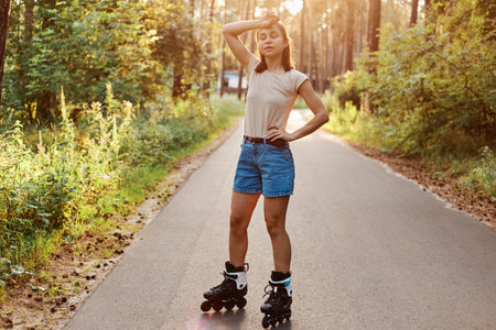 Tired female standing on road in summer park, being exhausted to roller skate, stops to have rest,...