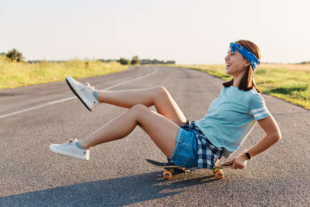 Side view of happy emotional woman sitting on skateboard on the street with raised legs, overjoyed female wearing casual clothing having fun outdoors, positive lifestyle.の写真素材
