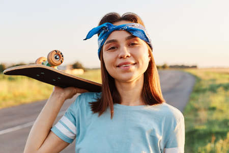 Portrait of beautiful brunette female wearing blue t shirt and hair band, holding skateboard over shoulder and looking at camera, enjoying skateboarding in summertime, active pastime.の写真素材
