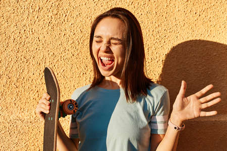 Photo of excited dark haired female wearing blue t shirt standing against yellow wall outdoor and screaming happily, holding longboard in hands, expressing happiness.の写真素材