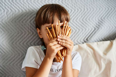 Little girl holding in hands and covering her face with biscuit sticks, dark haired unknown female child wearing white casual t shirt, sitting on sofa, looking through pretzels.の写真素材