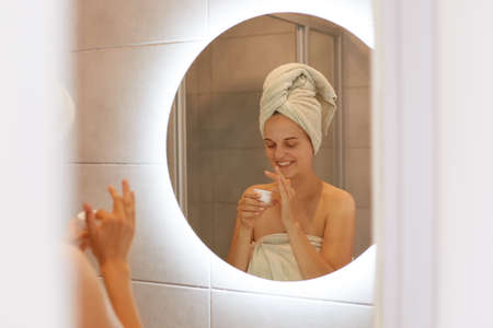 Portrait of woman being wrapped in white towel posing in bathroom in front of mirror open cream for...