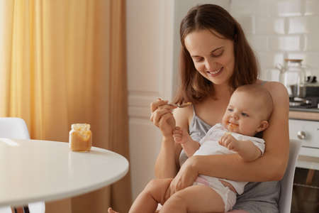 Indoor portrait of mother and child sitting in kitchen at table, woman with charming smile feeding baby with vegetable or fruit puree, complementary feeding of kid.の写真素材