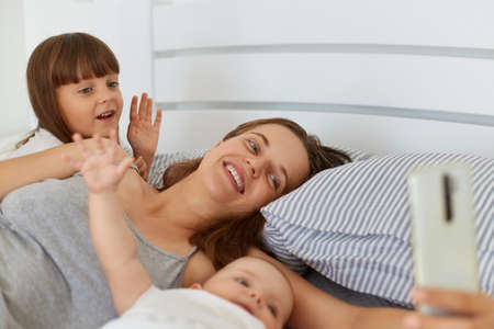 Happy positive female lying in bed with her children and holding smart phone in hands, people waving hands ans say hello, family blog, online conversation with followers.の写真素材