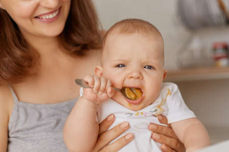 Indoor shot of happy smiling mother holding little child, cute baby trying eating fruit or vegetable...