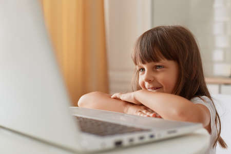Portrait of smiling small dark haired female child sitting at table in front of laptop computer and smiling, little preschooler girl watching cartoons or online lesson.の写真素材