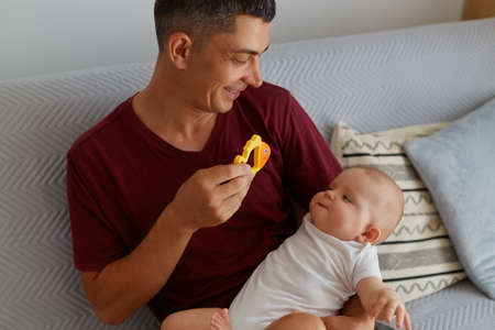 Happy father holding toy and playing with his baby boy or girl while sitting on sofa, smiling man wearing maroon t shirt showing to child orange fish, happy parenthood.の写真素材