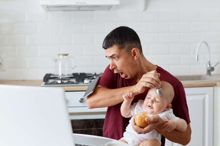 Shocked handsome man wearing maroon casual t shirt drops smart phone while sitting at table in kitchen and feeding little daughter or son, scared father with baby boy or girl.の写真素材