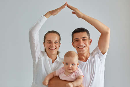 Indoor shot of happy positive family posing isolated light background, looking at camera, holding baby in hands, mom and dad making roof figure with hands arms over heads, safety.の写真素材