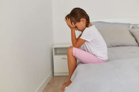 Side view portrait of sad female kid wearing white t shirt and pink short, dark haired child with braids sitting with head down, covering her face, being offended, expressing sadness.の写真素材