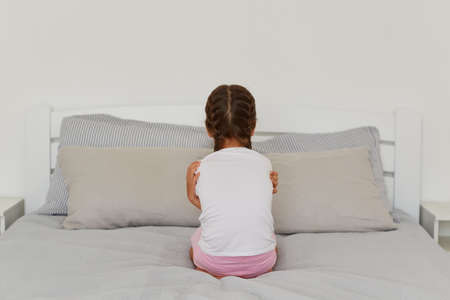 Back view portrait of little girl with dark hair and pigtails, siting backwards to camera, does not want talk with anybody, being offended, posing alone in her room.の写真素材