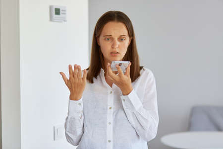 Portrait of confused puzzled female wearing white shirt posing at home with smart phone in hands,...