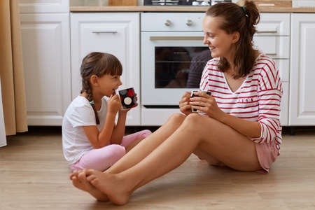 Full length view of happy mother drinking tea from cups with her smiling daughter, family sitting on kitchen floor, having breakfast and spending weekend together at homeの写真素材