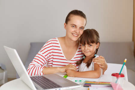 Indoor shot of happy positive woman with daughter, sitting at table with portable computer and...