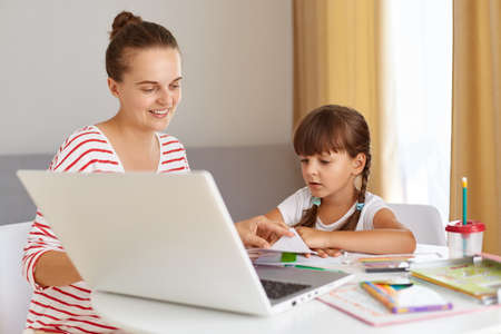 Portrait of happy positive female with daughter wearing casual attire, sitting at table against of...