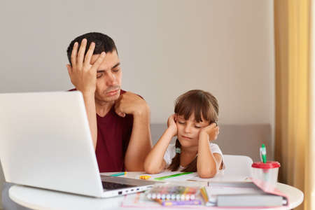 Portrait of sleeping daughter and father sitting at the desk while doing homework together, being...
