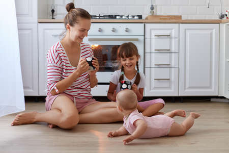 Full length portrait of happy mother wearing casual striped shirt sitting at floor and drinking hot coffee or tea while spending time with her daughters posing near woman.の写真素材