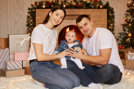 Smiling wife and husband with little toddle daughter sitting on the floor at home with xmas tree and...