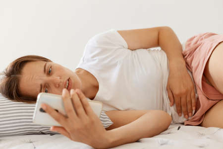 Sick woman with frowning face wearing white casual t shirt lying in bed, holding smart phone in hands, needs to call to doctor, suffering from terrible stomachache.の写真素材