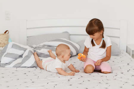Indoor shot of little smiling kids playing together sitting on bed, elder daughter showing small orange toy to her tiny sister, children posing on bed in light bedroom.の写真素材