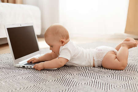 Indoor shot of baby playing with note book while lying on floor against the window, toddler wearing white t shirt and diaper, posing alone at home, happy childhood.の写真素材