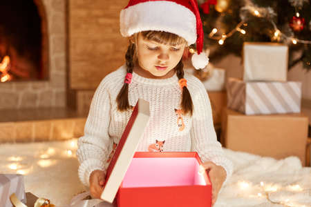 Little girl wearing white sweater and santa claus hat, opening present box with something glowing inside, posing in festive room with fireplace and Xmas tree.の写真素材